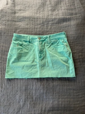 Turkos kort jeanskjol - Snygg mintgrön kort jeanskjol med rå kant nertill och klassiska fem fickor. Kjolen har bälteshällor och stängs med knapp och dragkedja framtill. Perfekt för dig som vill ha en fräsch och trendig look.