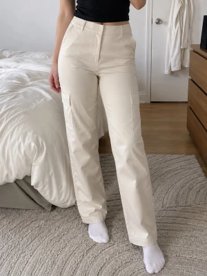 Beige cargobyxor med raka ben - Stilrena beige cargobyxor med raka ben och hög midja. Byxorna har klassiska cargofickor på sidorna och stängs med dragkedja och knapp. Perfekta för en trendig och avslappnad look. Materialet ser ut att vara bomull eller bomullsblandning.