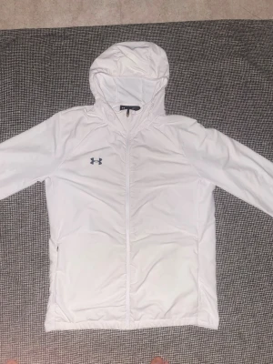 Vit vindjacka från Under Armour - Snygg vit vindjacka från Under Armour med huva och dragkedja framtill. Jackan har logga på bröstet och två sidofickor. Tillverkad i ett lätt och funktionellt material som passar perfekt för blåsiga dagar limited edition.