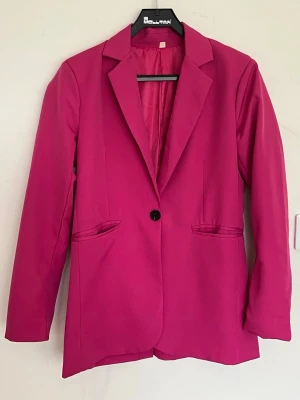 Fuchsia kavaj med enkel knapp - Stilren fuchsia kavaj med klassisk krage och en knapp framtill. Två fickor utan lock och helfodrad insida. Rak passform och långa ärmar. Perfekt för att addera färg till din outfit.