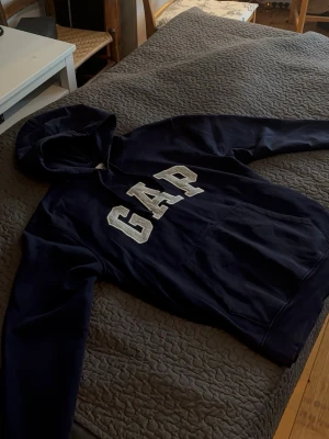 Mörkblå hoodie från GAP - Snygg mörkblå hoodie från GAP med stor vit logga på bröstet. Klassisk modell med huva och känguruficka framtill. Tillverkad i mjukt bomullsmaterial som är skönt att ha på sig. Perfekt för en avslappnad och casual stil.                 Inga som helst defekter eller märken/fläckar, som ny! Storlek L
