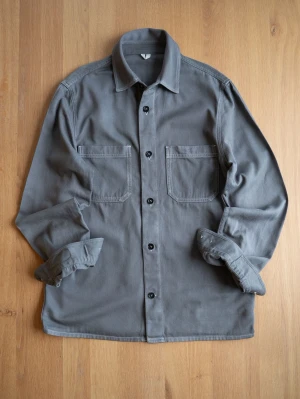 Grå Arket overshirt med fickor - Stilren grå Arket overshirt i kraftigt bomullstyg med två stora bröstfickor och svarta knappar framtill. Klassisk krage och raka ärmar som kan vikas upp för en avslappnad look. Perfekt lager-på-lager-plagg för dig som gillar minimalistisk stil.