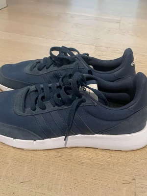 Mörkblå Adidas sneakers med vit sula - Snygga mörkblå sneakers från Adidas med klassiska tre ränder på sidan och vit, tjock sula. Skorna har snörning och är tillverkade i textil och syntetmaterial. Perfekta för en sportig och avslappnad stil.