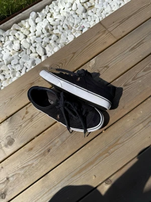 Polo Ralph Lauren sneakers - Snygga sneakers från Polo Ralph Lauren med klassisk låg profil och vit sula. Skorna har tygmaterial, svarta skosnören och den ikoniska broderade Polo-loggan i guld på sidan. Perfekta för en avslappnad och stilren look.