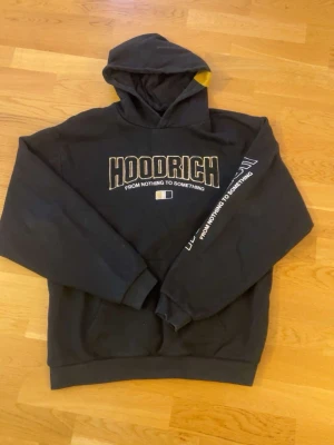 Svart HOODRICH hoodie - Svart hoodie från HOODRICH med stora tryck på både fram- och baksida samt ärm. Texten 'FROM NOTHING TO SOMETHING' och flera coola grafiska detaljer. Klassisk huva med gul detalj på insidan och ribbade muddar. Perfekt för dig som gillar streetwear och sportig stil.