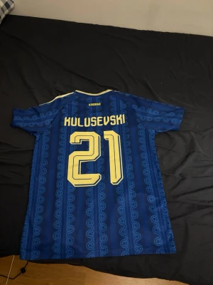 Blå Sverige fotbollströja Kulusevski - Säljer en blå fotbollströja från Adidas med gult tryck, nummer 21 och namnet Kulusevski på ryggen. Tröjan har korta ärmar, gul detalj vid kragen och ett mönster i tyget. Perfekt för dig som vill stötta Sverige på match!