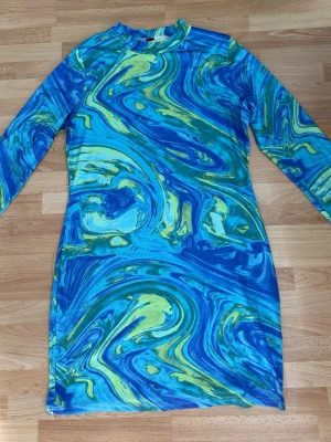 Blågrön mönstrad långärmad klänning - Cool långärmad klänning med swirl-mönster i blått, grönt och gult. Klänningen har en kroppsnära passform och rund halsringning. Perfekt för dig som vill sticka ut med färg och mönster. Sydde upp armarna till3/4. Lappar bortklippta. Använd en gång.