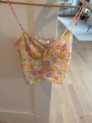 Blommig crop top från Bershka - Säljer en färgglad blommig linnetopp från Bershka med tunna axelband och söt knytning framtill. Toppen har ett somrigt mönster i orange, gul, lila och grön på vit botten. Perfekt för varma dagar och enkel att styla med jeans eller kjol.