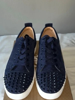 Mörkblå sneakers med nitar Louboutin - Säljer ett par mörkblå sneakers från Christian Louboutin med coola svarta nitar på tån och klassisk röd sula. Skorna har snörning och är tillverkade i ett tygmaterial som ger en lyxig känsla. Perfekta för dig som vill sticka ut med din stil.  Priset kan diskuteras