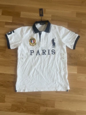 Vit pikétröja från Polo Ralph Lauren - Vit pikétröja från Polo Ralph Lauren med mörkblå detaljer, broderad logga och texten PARIS på bröstet. Klassisk krage med knappslå och emblem på vänster sida. Perfekt för en sportig och stilren look.