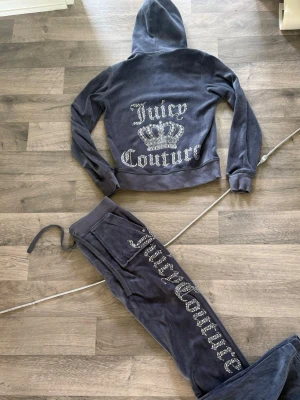 Juicy Couture grå velourset - Mysigt loungewear-set i grå velour från Juicy Couture. Setet består av en hoodie med dragkedja och fickor samt matchande byxor med dragsko. Båda plaggen har glittrande Juicy Couture-tryck och kronmotiv i strass på ryggen och benet.