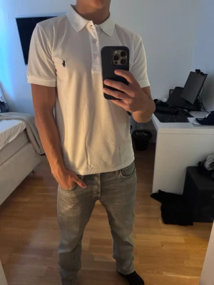 Vit pikétröja från Polo Ralph Lauren - Klassisk vit pikétröja från Polo Ralph Lauren med korta ärmar, krage och knappar framtill. Tröjan har den ikoniska broderade loggan på bröstet och är tillverkad i mjuk bomull. Perfekt för en clean och stilren look.