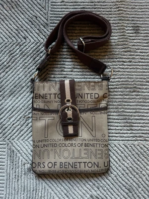 Beige axelrems väska United Colors of Benetton - Snygg beige axelrems väska från United Colors of Benetton med brunt och vitt mönsterband och logotyptryck över hela väskan. Justerbar axelrem i tyg och coolt metallspänne framtill. Perfekt för dig som gillar streetstyle och vill ha en unik accessoar. Jättefint skick! OBS: FRAKTAR INOM 24H!🩷