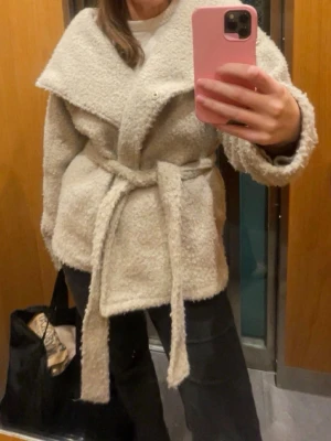 Beige teddykofta med knytskärp - Mysig beige cardiganjacka i teddy-material med bred krage och knytskärp i midjan. Jackan har en avslappnad passform och långa ärmar, perfekt för lager på lager. Passar dig som gillar en trendig och bekväm stil.