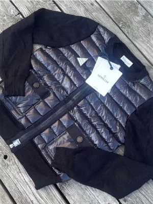 Svart pufferjacka från Moncler - Snygg svart pufferjacka från Moncler med glansig quiltad front och stickade ärmar samt muddar. Jackan har dragkedja framtill, två fickor med knapp och ribbade detaljer. Perfekt för dig som vill ha en trendig och bekväm jacka med exklusiv känsla.