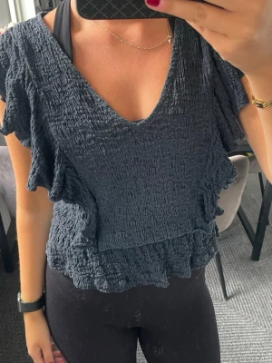 Mörkblå volangblus med v-ringning - Säljer en mörkblå blus med v-ringning och volangdetaljer längs ärmar och nederkant. Toppen har en skrynklig struktur och är kortare i modellen. Perfekt för dig som gillar en luftig och trendig stil.