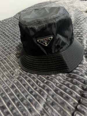 Svart bucket hat från Prada - Stilren svart bucket hat från Prada i glansigt nylonmaterial. Klassisk form med ikonisk metallogo framtill. 