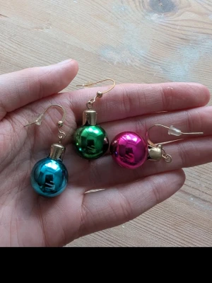Färgglada julkuleörhängen - Unika örhängen formade som små julkulor i metallicblått, grönt och rosa. Kulorna har en glansig yta och guldfärgade detaljer. Perfekta för dig som vill sticka ut och addera lite färg till din stil.
