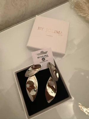 Silvriga statementörhängen By Jolima - Säljer ett par stora, silvriga statementörhängen från By Jolima. Örhängena är tillverkade i blankt rostfritt stål och har en modern, böljande form med snygg struktur på baksidan. Perfekta för dig som vill sticka ut och addera en cool touch till din look.