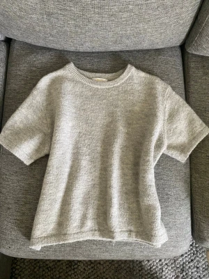 Ljusgrå stickad t-shirt - Kortärmad stickad t-shirt i ljusgrått med rund halsringning och avslappnad passform. Tröjan har ribbstickad kant vid halsen och är gjord i ett mjukt, mysigt material som passar perfekt till lager på lager. Enkel och stilren design utan synliga loggor.