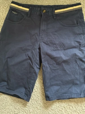 Marinblå shorts från Hackett London - Snygga marinblå shorts från Hackett London med klassisk femficksdesign och en beige randig detalj i midjan. Den är använd några fåtal gånger och köpte den för 1100 kr.