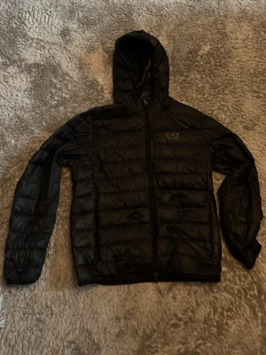 Svart pufferjacka från EA7 - Snygg svart pufferjacka från EA7 med huva och dragkedja framtill. Jackan har en stilren design med diskret logga på bröstet och quiltad struktur. Perfekt för kalla dagar när du vill hålla stilen och värmen.