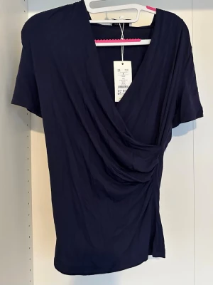Mörkblå omlottopp - Snygg mörkblå omlottopp med korta ärmar och djup v-ringning. Toppen har en draperad effekt framtill som ger en schysst siluett. Perfekt att styla med jeans eller kjol för en trendig look.