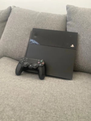 Playstation 4 (fat edition) - Säljer en svart PlayStation 4 från Sony med tillhörande DualShock 4-handkontroll. Konsolen har en stilren, rektangulär design i matt och blank plast. Perfekt för gaming och streaming, med flera anslutningsmöjligheter på baksidan.