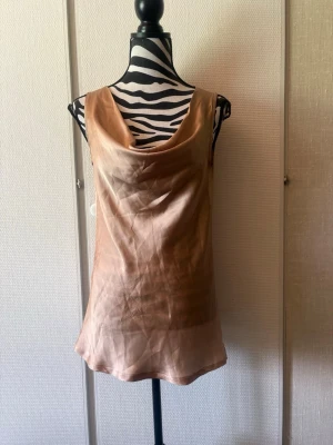 Champagnefärgad linnetopp i satin - Snygg ärmlös linnetopp i champagnefärgad satin med draperad urringning. Toppen har en slät baksida och ett lätt glansigt material som ger en lyxig känsla. Perfekt att styla med jeans eller kjol för en trendig look.