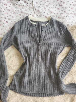 Grå ribbstickad tröja med knappar - Köpt från simple retro, super bra kvalite! Inga defekter💖Mysig grå ribbstickad tröja med långa ärmar och knappslå framtill. Tröjan har en vit kontrastkant vid halsen och är figurnära i modellen. Perfekt för lager på lager och enkel att styla till olika outfits.