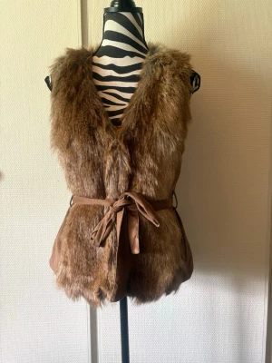 Brun pälsväst med knytband - Snygg brun pälsväst i fuskpäls med mjukt material och ett matchande knytband i midjan. Västen har djup V-ringning och är ärmlös, vilket ger en trendig och avslappnad look. Perfekt att slänga över en hoodie eller långärmad tröja för extra stilpoäng.