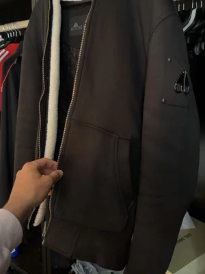 Svart pufferjacka Moose Knuckles - Svart pufferjacka från Moose Knuckles med dragkedja, stora fickor framtill och vit fuskpäls på insidan. Jackan har en klassisk pufferlook och en metallogo på ärmen. Perfekt för kalla vinterdagar och riktigt varm.