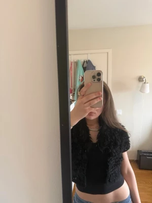 Svart pälsväst från Clara - Snygg svart pälsväst från Clara med fluffig, lockig fuskpäls och virkade detaljer. Kort modell som slutar vid midjan och har dekorativa bollar framtill. Perfekt för att lyfta din outfit med en trendig touch.
