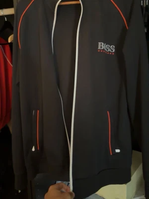 Svart ziptröja från Boss med röda detaljer - Snygg svart ziptröja från Boss med röda och vita detaljer. Tröjan har dragkedja framtill, två fickor och logga på bröstet. Perfekt för en sportig och stilren look.
