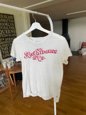 Vit Levi's t-shirt med rosa tryck - Säljer en vit t-shirt från Levi's med korta ärmar och ett stort rosa 'Levi Strauss & Co.' tryck på bröstet. T-shirten har en klassisk passform och är gjord i mjuk bomull, perfekt för en avslappnad och stilren look.