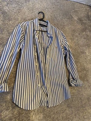 Randig skjorta från Polo Ralph Lauren - Säljer en blåvit randig skjorta från Polo Ralph Lauren i slim fit. Skjortan sitter som den ska, alltså inte för långt ned och allmänt super fräsch inför sommaren! Tillverkad i mjuk bomull och har den ikoniska lilla logobroderingen på bröstet.