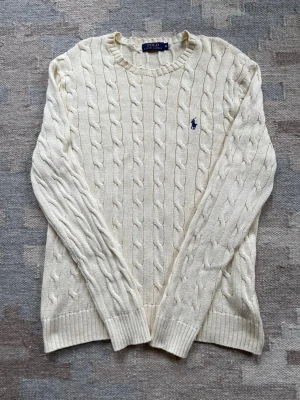 Vit kabelstickad tröja Polo Ralph Lauren - Kabelstickad vit tröja från Polo Ralph Lauren med rund halsringning och klassisk blå logga broderad på bröstet. Tröjan har långa ärmar och ribbade muddar vid ärmslut och nederkant. Perfekt för en clean och stilren look.
