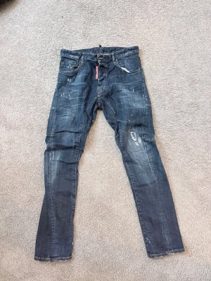 Blå distressed jeans från Dsquared2 - Säljer ett par blå jeans från Dsquared2 med slim fit och coola slitningar, färgstänk och patch-detaljer. Jeansen har klassisk femficksmodell, knappgylf och är gjorda i bomull med lite stretch. Perfekta för dig som gillar en edgy streetstil. Storlek 46 ny pris + 2500 kr säljer för endast 1110