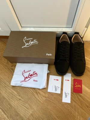Svarta sneakers från Christian Louboutin - Snygga svarta sneakers från Christian Louboutin med klassisk röd sula och svarta skosnören. Skorna har en stilren design i mocka och läder, med lågt skaft och diskreta detaljer. Perfekta för dig som vill ha något exklusivt och trendigt i garderoben.