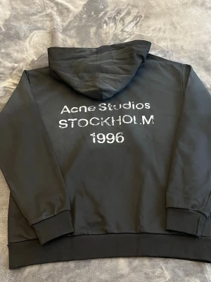 Svart hoodie Acne Studios Stockholm - Svart hoodie från Acne Studios med tryck i vitt på ryggen där det står 'Acne Studios STOCKHOLM 1996'. Klassisk passform med huva och muddar vid ärmslut och nederkant. Perfekt för en stilren och urban look.