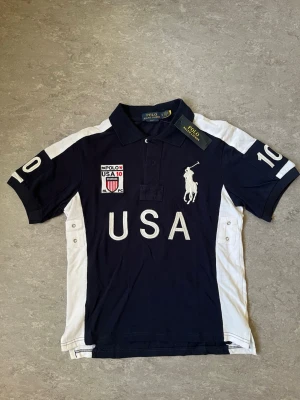 Polo Ralph Lauren USA piké - Marinblå och vit t-shirt från Polo Ralph Lauren med stora broderade detaljer, USA-tryck och logga på bröstet. Klassisk pikékrage och korta ärmar. Snygga kontrastpaneler i vitt på sidorna och ärmarna. Tillverkad i bomull för skön känsla.