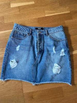 Blå jeanskjol från Bik Bok - Säljer en blå jeanskjol från Bik Bok med slitna detaljer och rå kant nertill. Kjolen har klassisk femficksdesign och knappgylf. Perfekt för dig som gillar en avslappnad och trendig stil.