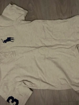 Beige pikétröja från Polo Ralph Lauren - Klassisk beige pikétröja från Polo Ralph Lauren med blå logga på bröstet och siffran 3 på ärmen. Tröjan har krage, knappar vid halsen och är i modellen Classic Fit. Perfekt till en avslappnad men ändå stilren look.