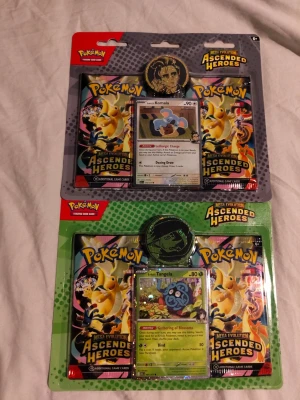 Pokémon Ascended Heroes boosterpaket - Två Pokémon Ascended Heroes boosterpaket med samlarkort och mynt. Ena paketet innehåller ett Komala-kort och det andra ett Tangela-kort. Båda paketen har färgglada förpackningar och inkluderar flera booster packs samt ett samlarmynt.