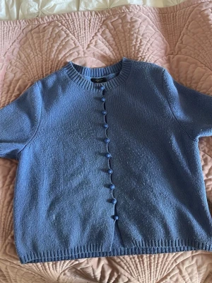 Blå stickad kofta Massimo Dutti - Säljer en blå stickad kofta från Massimo Dutti med korta ärmar och rund hals. Koftan har matchande blå knappar framtill och ribbstickade kanter vid hals, ärmslut och nederkant. Perfekt att slänga över en topp för en chill look.( PRIS KAN DISKUTERAS)