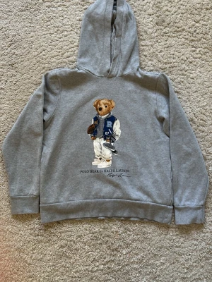 Grå hoodie Polo Bear Ralph Lauren - (Storlek 14-16 L)Snygg grå hoodie från Polo Ralph Lauren med den ikoniska Polo Bear-trycket på bröstet. Klassisk huva och ribbade muddar. Tillverkad i mjuk bomullsblandning för skön komfort. Perfekt för en avslappnad och trendig stil.
