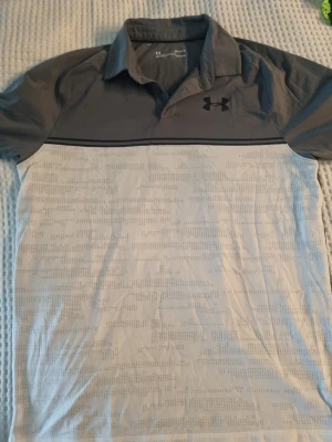 Grå och vit pikétröja från Under Armour - Snygg pikétröja från Under Armour i grått och vitt med diskret mönster på nederdelen. Tröjan har klassisk krage, knappar och logga på bröstet. 