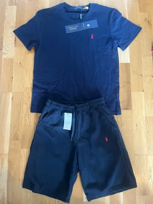 Mörkblått set från Polo Ralph Lauren - Snyggt mörkblått set från Polo Ralph Lauren med t-shirt och shorts. Båda plaggen har den klassiska röda logotypen broderad på bröstet och benet. T-shirten har rund hals och shortsen har resår i midjan med dragsko. Perfekt för en avslappnad stil.