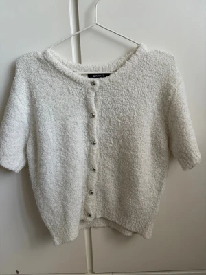Vit kortärmad kofta från Gina Tricot - Mysig vit kofta från Gina Tricot med korta ärmar och dekorativa pärlknappar framtill. Tillverkad i ett fluffigt, bouclé-liknande material som ger en trendig och avslappnad look. Perfekt att styla över ett linne eller klänning.