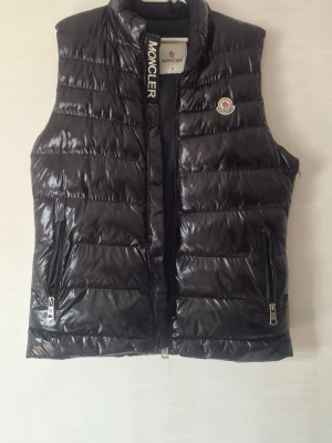 Svart dunväst från Moncler - Snygg svart dunväst från Moncler med glansig finish och klassisk quiltad design. Västen har hög krage, två dragkedjeförsedda fickor och Moncler-logga på bröstet. Perfekt för lager-på-lager och streetstyle.